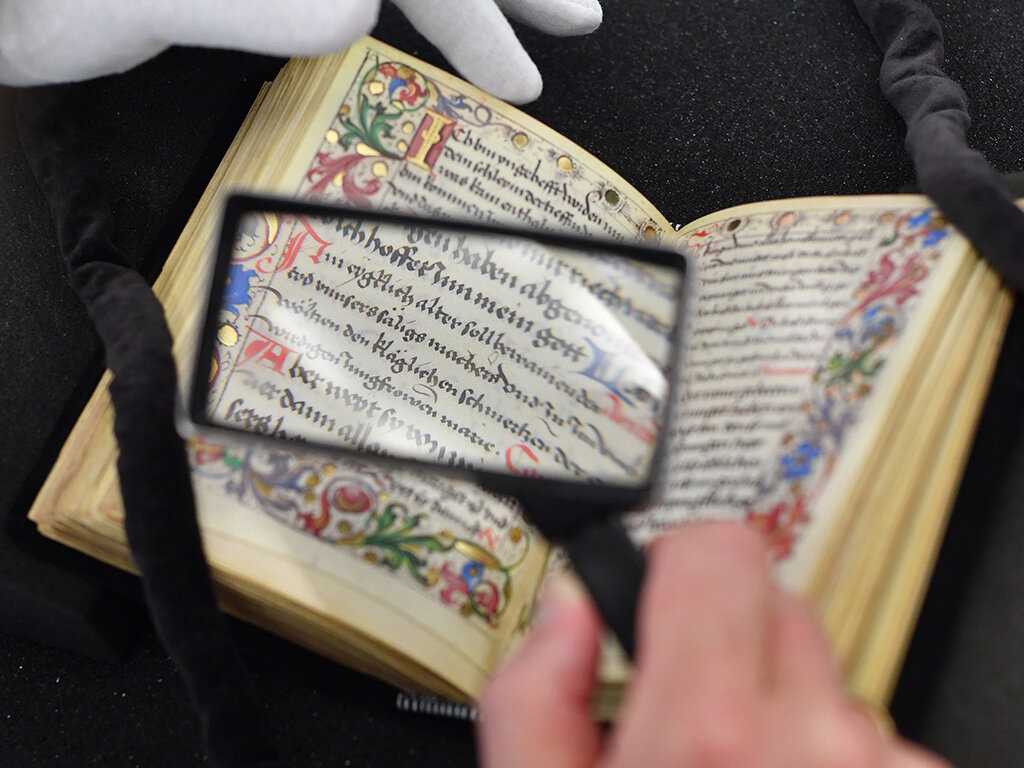Zu sehen ist ein historisches Buch, welches durch eine Lupe betrachtet wird. Der Bildausschnitt offeriert nur das Buch, eine Hand mit einer Lupe und eine weitere mit einem weißen Handschuh. 