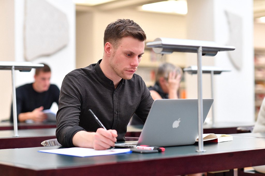 Das Bild zeigt einen Nutzer an einem der Arbeitsplätze im Lesesaal der Badischen Landesbibliothek. Er blickt in ein vor ihm auf dem Tisch stehendes Laptop und hält mit der rechten Hand einen Stift bereit, um sich Notizen zu machen.