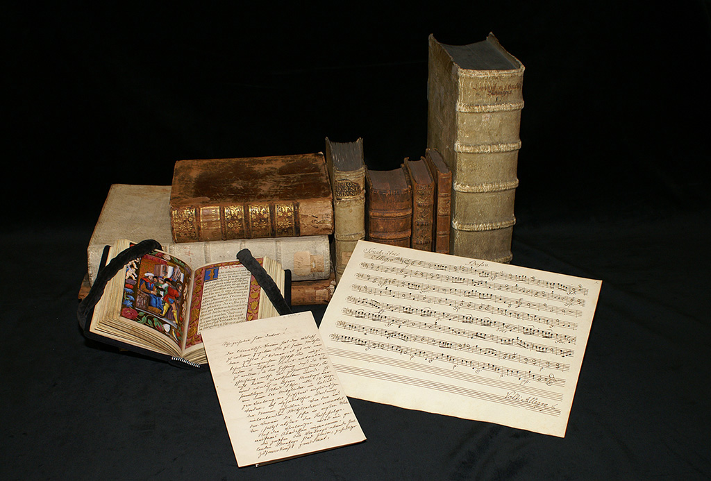Auf dem Bild sieht man ausgewählte Sammlungsbestände der Badischen Landesbibliothek, aufgeschlagen im Vordergrund eine mittelalterlichen Handschrift und ein Notenblatt