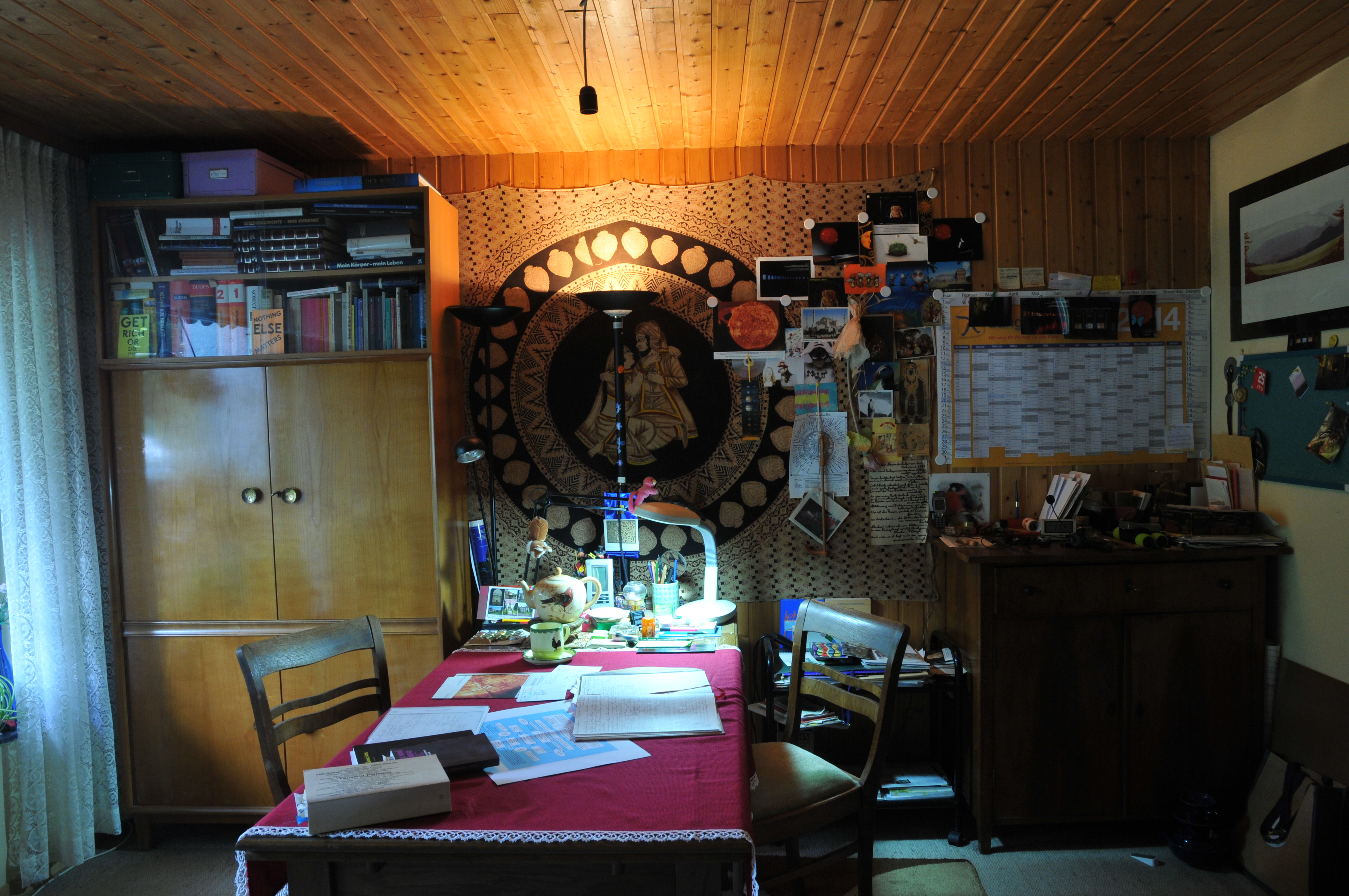Fotografie des Arbeitszimmers von Krebs, in der Mitte steht ein Tisch mit roter Decke, die Wände sind mit Holz vertäfelt, über sind Bücher, Bilder und Notizen.