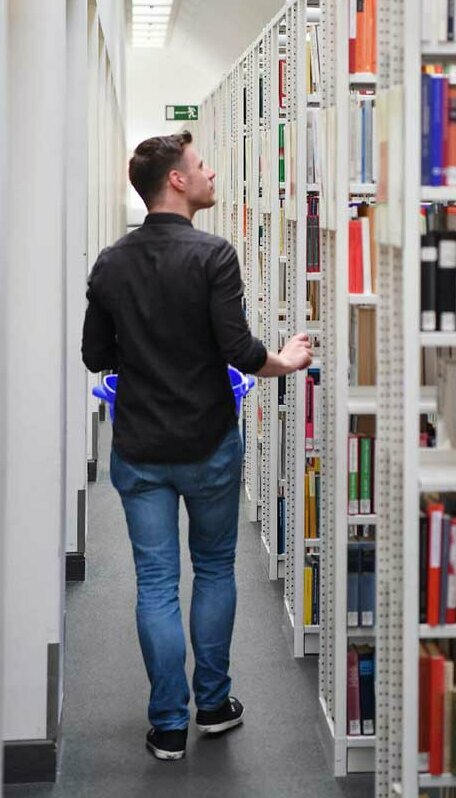 © Foto: Uli Deck Das Bild zeigt in Rückenansicht einen Nutzer, der mit einem Bücherkorb durch die Gänge des offenen Magazins läuft.