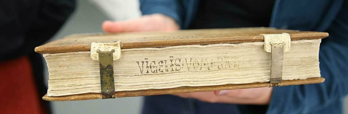 Hände halten den Buchschnitt der Handschrift in die Kamera. Die Handschrift hat zwei Beschläge, auf dem Schnitt steht schlecht leserlich etwas geschrieben.