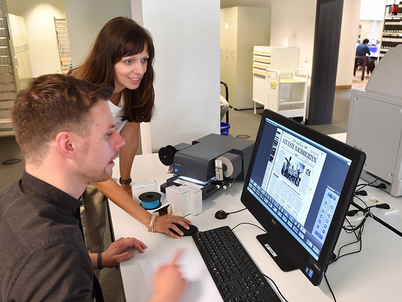 Eine Mitarbeiterin erklärt einem Benutzer an einem PC die Nutzung der Mikrofilmscanner.