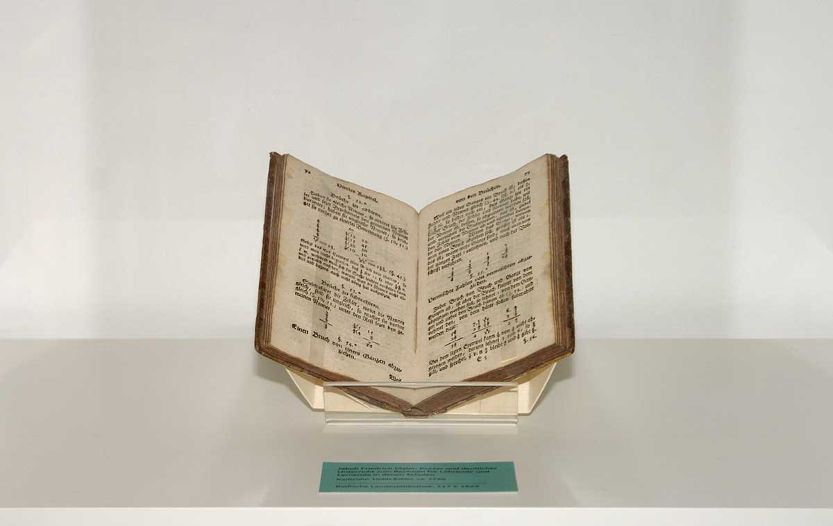 © Foto: Badische Landesbibliothek Karlsruhe Aufgeschlagen in einer Vitrine steht Jakob Friedrich Maler's Schulbuch. Auf der Doppelseite werden mathematische Brüche behandelt.