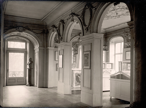 Schwarz-weiß Fotografie des ehemaligen Bibliothekssaal im Schloss. Prunkvoller Raum mit Büsten, Schaukästen und Bildern an den Wänden.