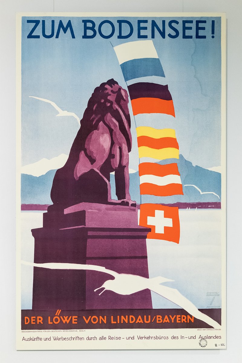 Plakat mit einer grafischen Abbildung des bayerischen Löwen von Lindau mit der Aufschrift „Zum Bodensee!“ Seitlich sind diverse Flaggen abgebildet.
