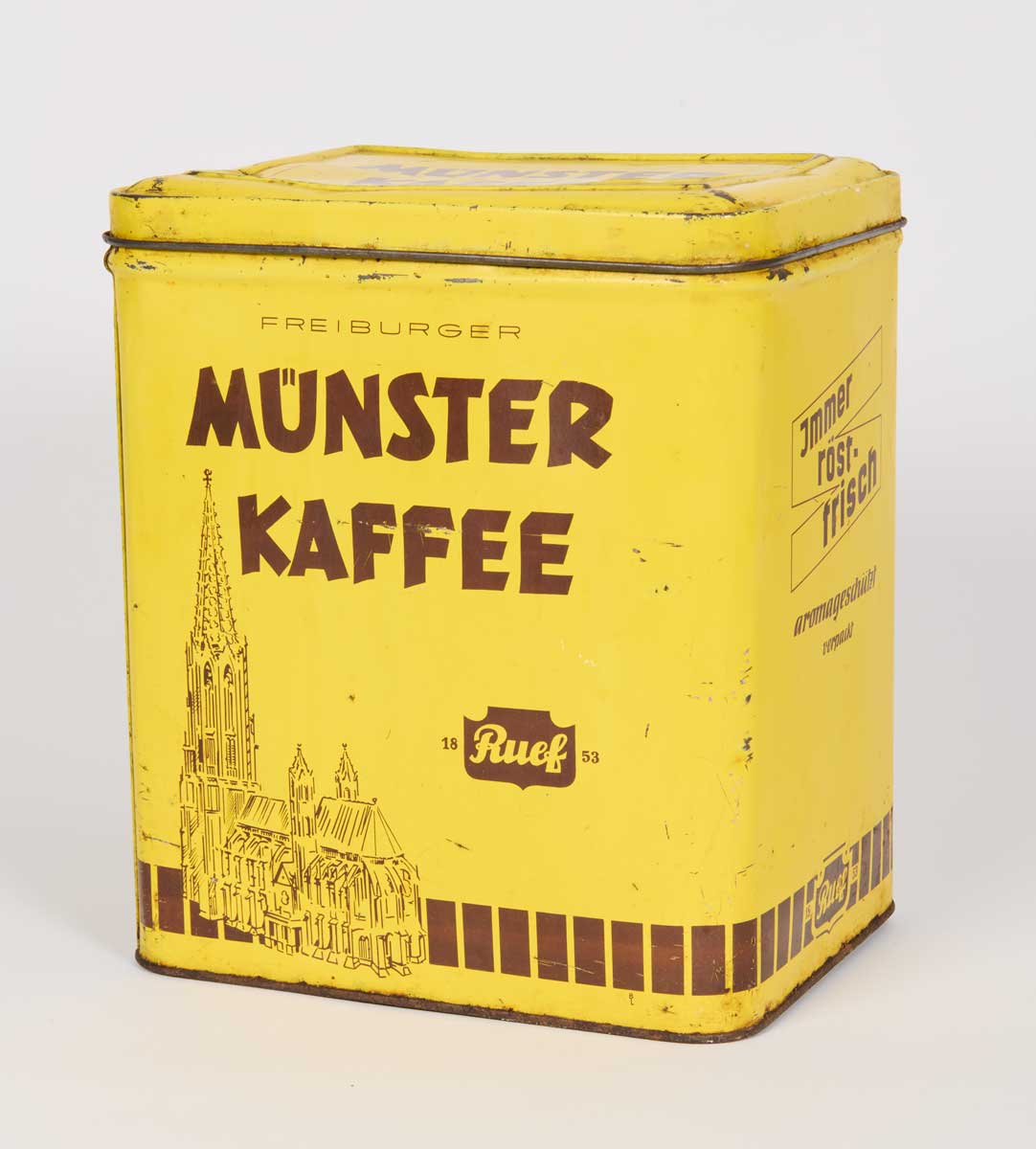 Das Foto zeigt eine gelbe Blechdose zur Aufbewahrung von Kaffee. Vorne ist Freiburger Münster als Strichzeichnung in brauner Farbe abgebildet. Darüber steht die Marke „Freiburger Münster-Kaffee“. Auf der rechten Seite ist folgender Text zu erkennen: „Immer röstfrisch, aromageschützt verpackt“.