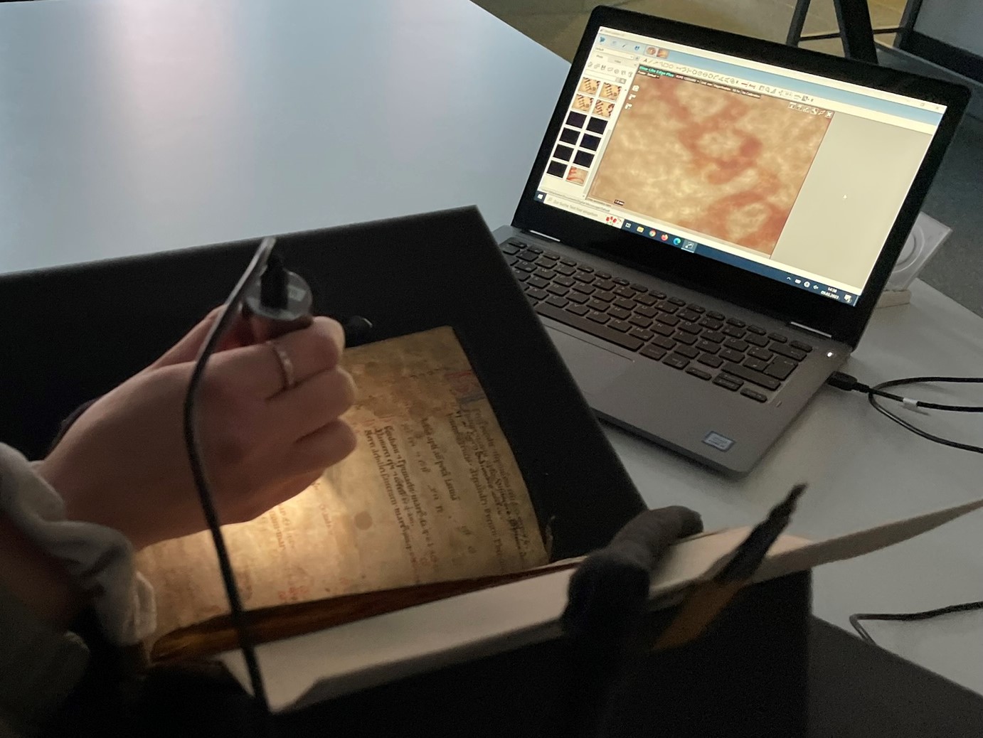 Zu sehen ist das Digitalmikroskop, das bei einem alten gedruckten Buch eingesetzt wird und auf dem Laptop im Hintergrund einen stark vergrößerten Ausschnitt zeigt.