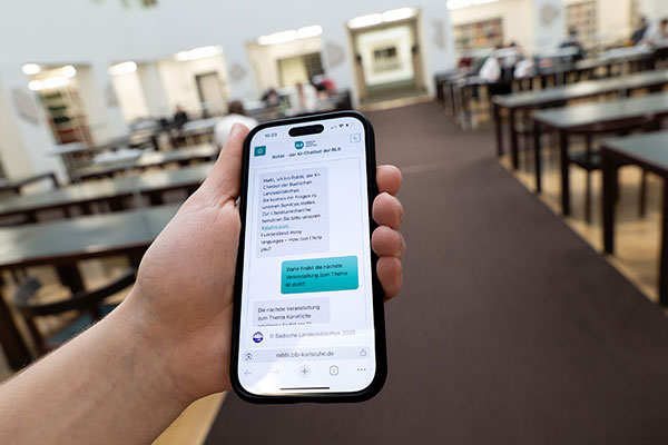 Das Foto zeigt eine Hand, die ein Mobiltelefon umgreift. Auf dem Display ist der Chatverlauf mit Chatbot „Robbi“ zu sehen. Im Hintergrund sind verschwommen die Tische des Lesesaals abgebildet.